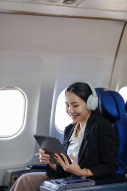 Bir kadın kucağında tabletle uçakta oturuyor. Kulaklık takıyor ve uçuşun tadını çıkarıyor.
