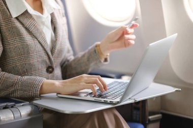 Önünde dizüstü bilgisayarı açık bir kadın uçakta oturuyor. Klavyeye yazı yazıyor ve elinde bir kredi kartı tutuyor. Verimlilik ve çalışma kavramı