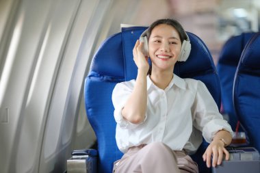 Kulaklık takan bir kadın mavi bir uçak koltuğunda oturuyor. Gülümsüyor ve uçakta eğleniyor.