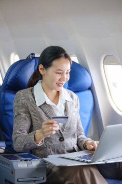 Bir kadın uçakta oturuyor ve biletini ödemek için dizüstü bilgisayar kullanıyor. Gülümsüyor ve uçakta eğleniyor.