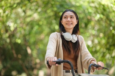 Parkta bisiklet süren bir kadın. Kulaklık takıyor ve gülümsüyor. Sahne huzurlu ve rahatlatıcı.