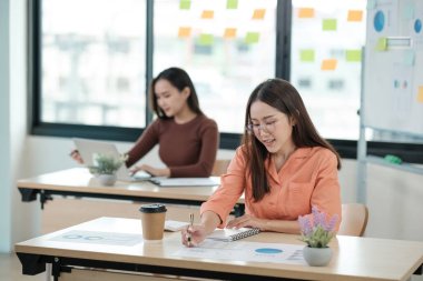 İki kadın masalarında oturuyor, biri deftere yazı yazıyor. Diğer kadın dizüstü bilgisayarda yazıyor. Sahne, insanların görevlerine odaklandıkları bir çalışma ortamı öneriyor.