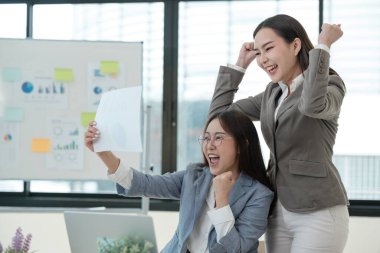 Takım elbiseli iki kadın zaferi kutluyor. Bir parça kağıt tutuyorlar ve gülümsüyorlar.