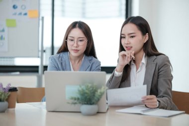 İki kadın bir dizüstü bilgisayar ve bir yığın kağıtla bir masada oturuyorlar. Laptop ekranına bakıyorlar ve bir şeyi tartışıyorlar. Sahne odaklı ve ciddi.