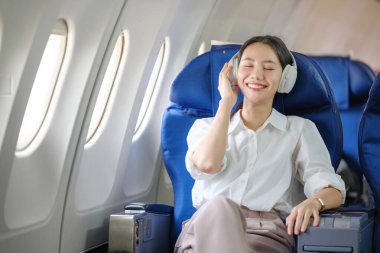 Bir kadın uçak koltuğunda oturmuş kulaklıkları takmış, gülümsüyor ve mutlu görünüyor. O müzik dinliyor ve uçuşunun tadını çıkarıyor.