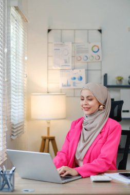 Pembe ceketli bir kadın önünde dizüstü bilgisayarla bir masada oturuyor. Gülümsüyor ve işinden zevk alıyor.