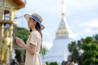 Hasır şapkalı ve beyaz gömlekli bir kadın bir binanın fotoğrafını çekiyor. Bina bir pagoda ve kadının elinde bir cep telefonu var.