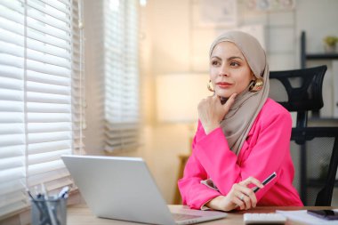Pembe takım elbiseli bir kadın dizüstü bilgisayarı ve kalemi olan bir masada oturuyor. Bir şey düşünüyor ya da düşünüyor.