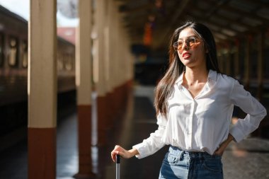 Güneş gözlüğü ve beyaz gömlek giyen bir kadın tren istasyonunda duruyor. Elinde bir bavul tutuyor ve bir tren bekliyor. Seyahat ve beklenti kavramı