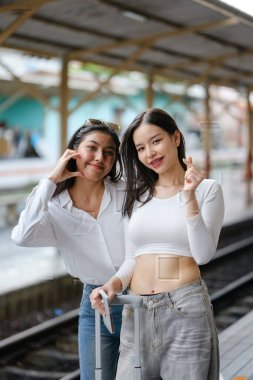 İki kadın cep telefonlarıyla fotoğraf çektirmek için poz veriyor. İçlerinden biri bir bavul tutuyor.