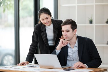 Bir kadın ve bir erkek birlikte dizüstü bilgisayara bakıyorlar. Kadın takım elbise giyiyor, erkek de takım elbise ve kravat takıyor. İkisi de dizüstü bilgisayara odaklanmış.