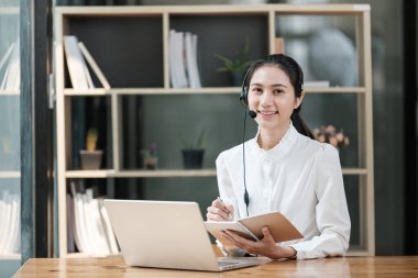Masasında dizüstü bilgisayarı ve not defteri olan bir kadın oturuyor. Gülümsüyor ve mutlu.