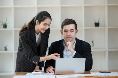 Bir kadın ve bir adam bir masada oturmuş laptopa bakıyorlar. Kadın ekrandaki bir şeyi işaret ediyor. Sahne profesyonel ve odaklı.