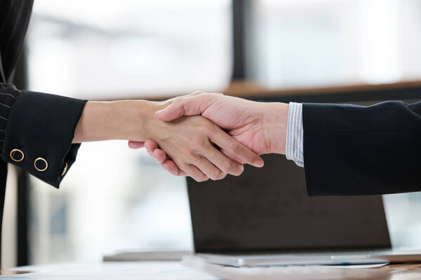 Два человека пожимают друг другу руки в деловой обстановке. На мужчине костюм, а на женщине чёрное платье. Перед ними ноутбук на столе.