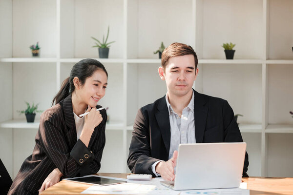 A man and a woman are sitting at a desk with a laptop open. The man is looking at the laptop while the woman looks at him. The scene suggests a professional setting where the man