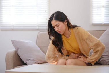 A woman is sitting on a couch with a stomach ache. She is wearing a yellow sweater and has a stomach ache clipart