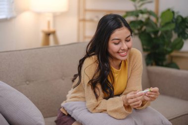 Elinde hamilelik testiyle kanepede oturan bir kadın. Gülümsüyor ve sonuç onu mutlu ediyor.