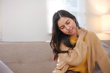 Boyun yaralanması olan bir kadın kanepede oturuyor. Sarı bir kazak giyiyor ve başını yana yatırmış.