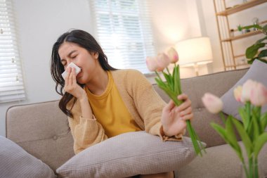 Bir kadın elinde mendille kanepede oturuyor. Burnunu çekiyor ve elinde bir buket çiçek tutuyor.
