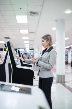 Bir kadın makinenin önünde duruyor ve telefonuna bakıyor. Gri bir ceket ve siyah pantolon giyiyor.