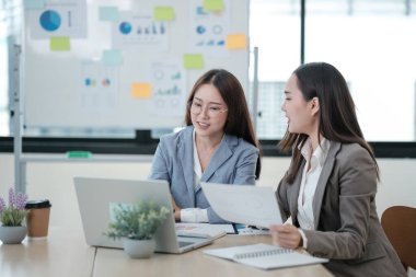 Önlerinde bir dizüstü bilgisayar ve kağıtlarla bir masada oturan iki kadın. Önemli bir şeyi tartışıyorlar, ve ifadeleri onların sohbete dahil olduğunu gösteriyor.