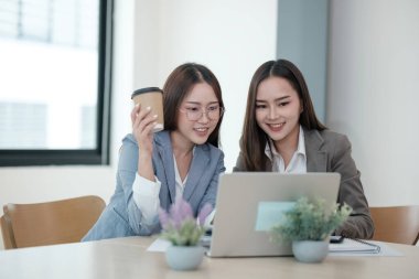 İki kadın bir masada oturuyor, biri bir fincan kahve tutuyor. Dizüstü bilgisayara bakıyorlar, muhtemelen iş ya da proje tartışıyorlar. Atmosfer profesyonel ve odaklanmış görünüyor.