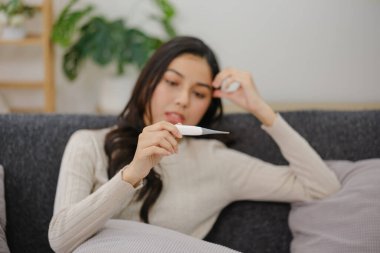 Bir kadın elinde termometreyle kanepede oturuyor. Rahatsızlık ya da hastalık durumunda.