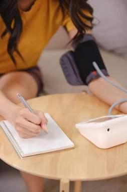 Kan basıncı monitörüyle masada oturan bir kadın deftere yazı yazıyor. Kadın tansiyonunu ölçerken odaklanma ve kararlılık kavramı