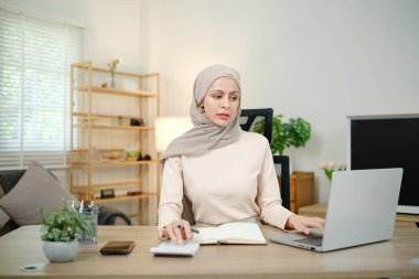 Başörtüsü takan bir kadın dizüstü bilgisayarı ve not defteri olan bir masada oturuyor. O, dizüstü bilgisayarda yazıyor ve deftere yazıyor. Odaklanma ve üretkenlik kavramı