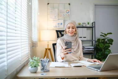 Başörtüsü takan bir kadın dizüstü bilgisayarı ve cep telefonu olan bir masada oturuyor. Gülümsüyor ve cep telefonuyla kendi fotoğrafını çekiyor.
