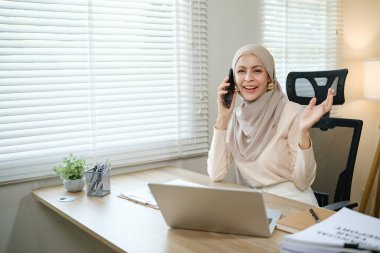 Başörtüsü takan bir kadın masada otururken cep telefonuyla konuşuyor. Gülümsüyor ve mutlu.