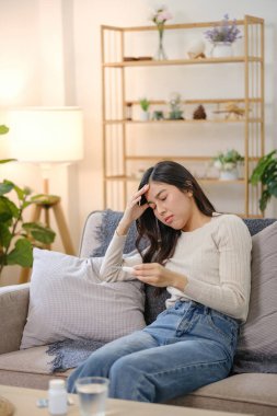 Kafası elinde bir kanepede oturan bir kadın. Elinde bir ilaç şişesi var ve acı çekiyor.