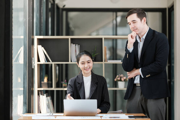 A man and a woman are standing in front of a desk with a laptop. The scene seems to be a work environment where the man and woman are collaborating on a project
