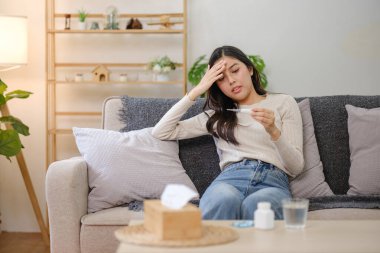 Bir kadın elinde termometreyle kanepede oturuyor. Alnına dayamış ve acı çekiyor.