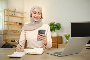 Başörtüsü takan bir kadın dizüstü bilgisayarı ve cep telefonu olan bir masada oturuyor. Gülümsüyor ve cep telefonuna bakıyor.