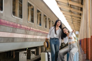 İki kadın bir tren peronunda bavullarıyla oturuyorlar. İçlerinden biri şapka tutuyor.