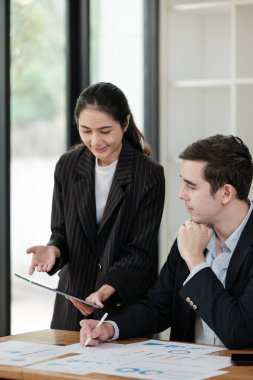 Bir kadın ve bir erkek önlerinde kağıtlarla bir masada oturuyorlar. Kadın kağıtlardaki bir şeyi işaret ederken, adam da ona bakıyor. Sahne profesyonel ve odaklı.