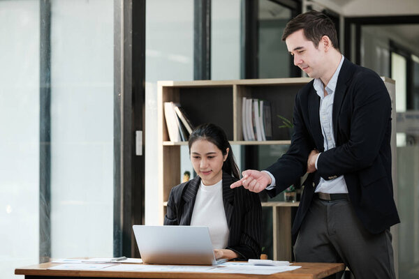 A man and a woman are sitting at a desk with a laptop. The man is pointing at something on the laptop screen