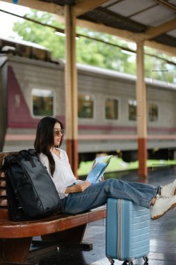Bir kadın bavul ve sırt çantasıyla bankta oturuyor. O bir kitap okuyor.