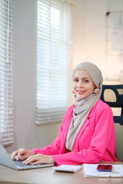 Pembe ceketli bir kadın elinde dizüstü bilgisayarla masada oturuyor. Gülümsüyor ve çalışıyor.