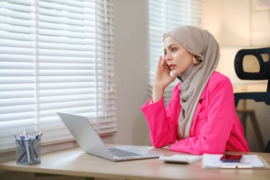 Pembe ceketli bir kadın masada otururken cep telefonuyla konuşuyor. O bir eşarp takıyor ve önünde bir laptop var.