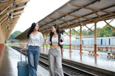 İki kadın bir tren platformunda bavullarıyla yürüyorlar. Manzara rahat ve rahat, çünkü kadınlar rahat giyinmiş ve yolculuklarının tadını çıkarıyorlar.