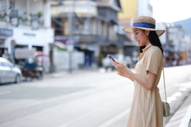 Bir kadın kaldırımda duruyor, cep telefonuna bakıyor. Hasır şapka ve bronz bir elbise giyiyor.