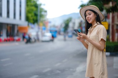 Sarı elbiseli bir kadın elinde cep telefonuyla caddede duruyor. Gülümsüyor ve eğleniyor.