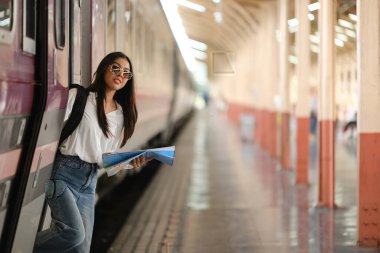 Bir kadın elinde bir kitapla tren peronunda duruyor. O, güneş gözlüğü ve beyaz bir gömlek giyiyor.