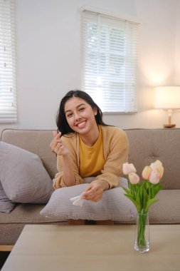 Bir kadın önünde bir vazo çiçekle bir kanepede oturuyor. Gülümsüyor ve elinde bir mendil tutuyor.