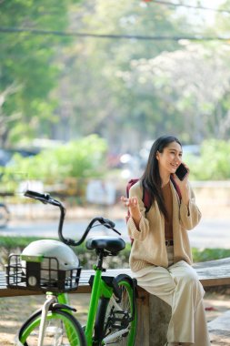 Yeşil bir bisikletin yanında bir kadın bankta oturuyor. Cep telefonuyla konuşuyor.