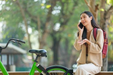 Yeşil bir bisikletin yanında bir kadın bankta oturuyor. Cep telefonuyla konuşuyor.