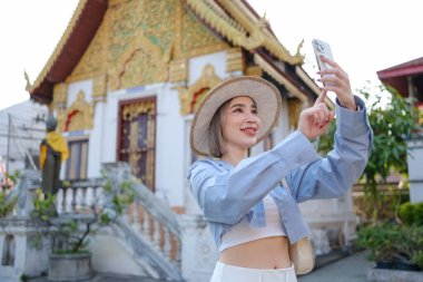 Hasır şapkalı bir kadın bir binanın fotoğrafını çekiyor. Bina süslenmiş ve çok fazla detay var. Kadın gülümsüyor ve eğleniyor gibi görünüyor.