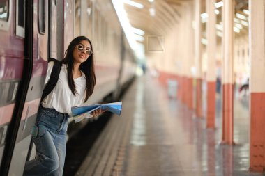 Bir kadın elinde bir kitapla tren peronunda duruyor. O, güneş gözlüğü ve beyaz bir gömlek giyiyor.